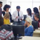 group of older students standing around, listening intently to a faculty member who wearing a white shirt and tie in a computer room. The faculty member's head is down as he's pointing to a piece of paper in his hand,